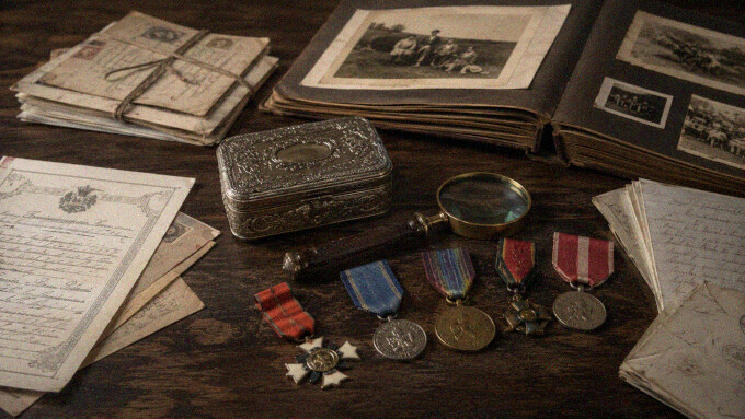 Historische Orden und Ehrenzeichen, alte Urkunden, Feldpostbriefe, geöffnetes Fotoalbum, Metallkassette und Lupe auf einem dunklen Holztisch.